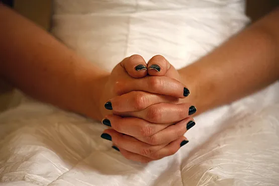 Nærbillede af en kvindes hænder med mørk neglelak, foldet i skødet, iført en hvid kjole eller brudekjole.