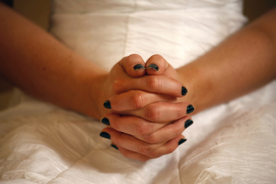 Nærbillede af en kvindes hænder med mørk neglelak, foldet i skødet, iført en hvid kjole eller brudekjole.