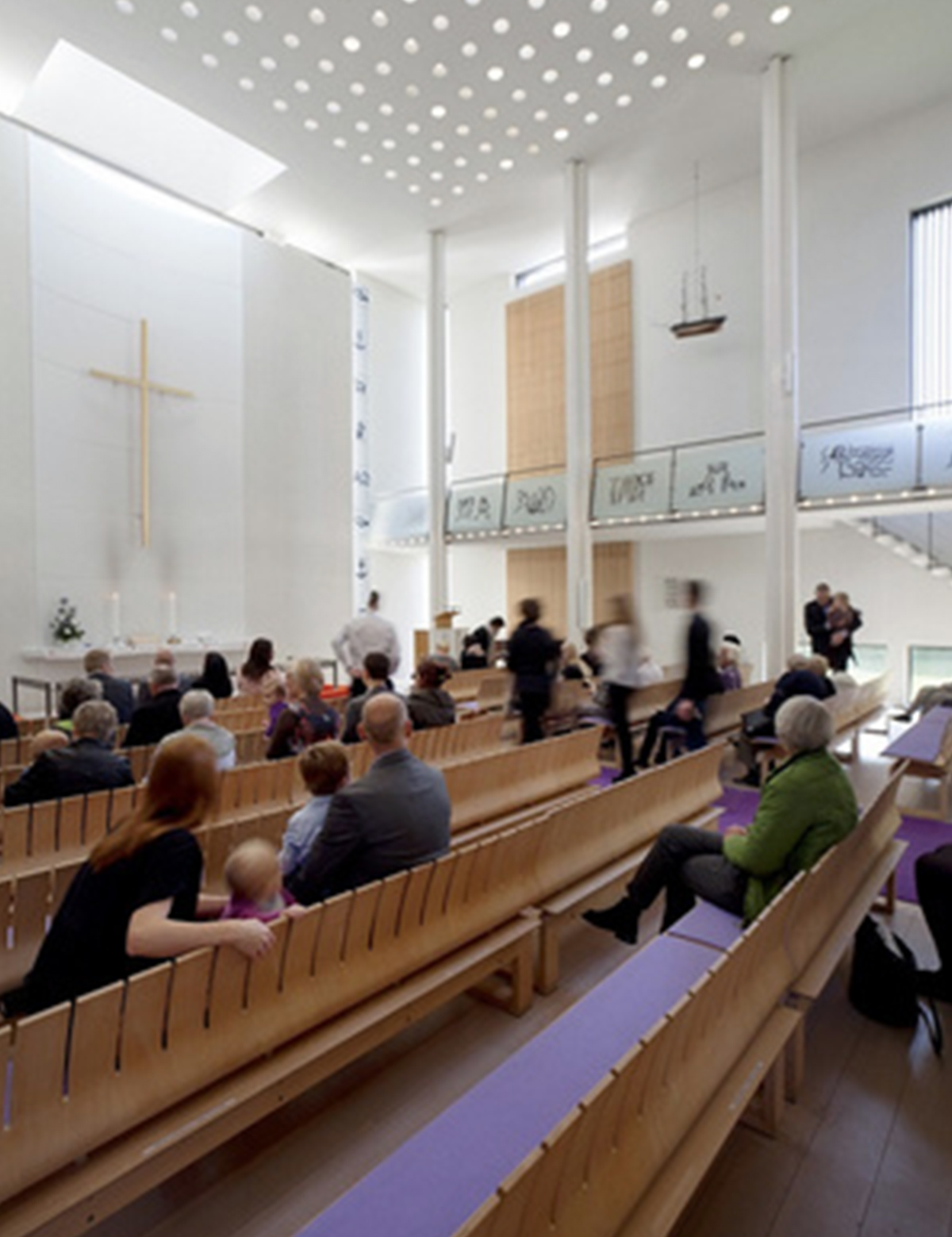 Lyst, moderne kirkeinteriør med folk i træbænke, et kors på væggen og et ophængt skib.