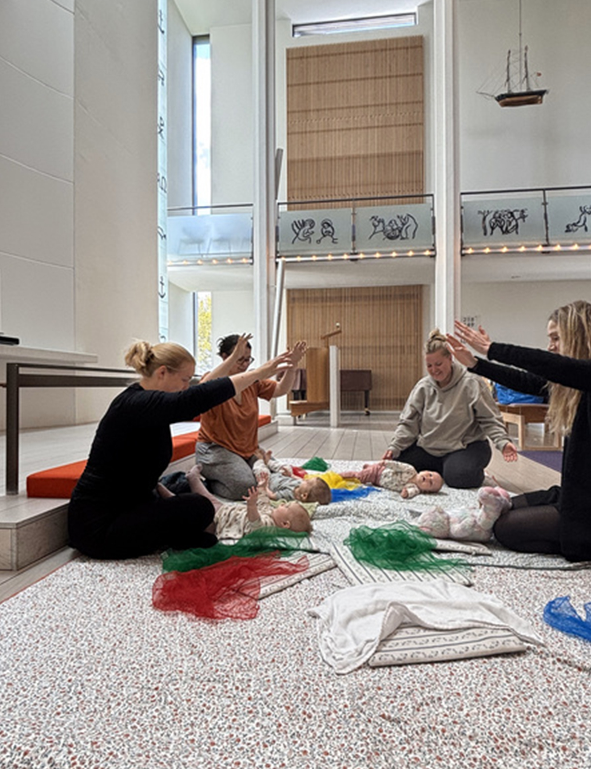 Gruppe af voksne og babyer, der sidder på gulvet i en kirke og deltager i en musikaktivitet med farvede tørklæder og gestusser.