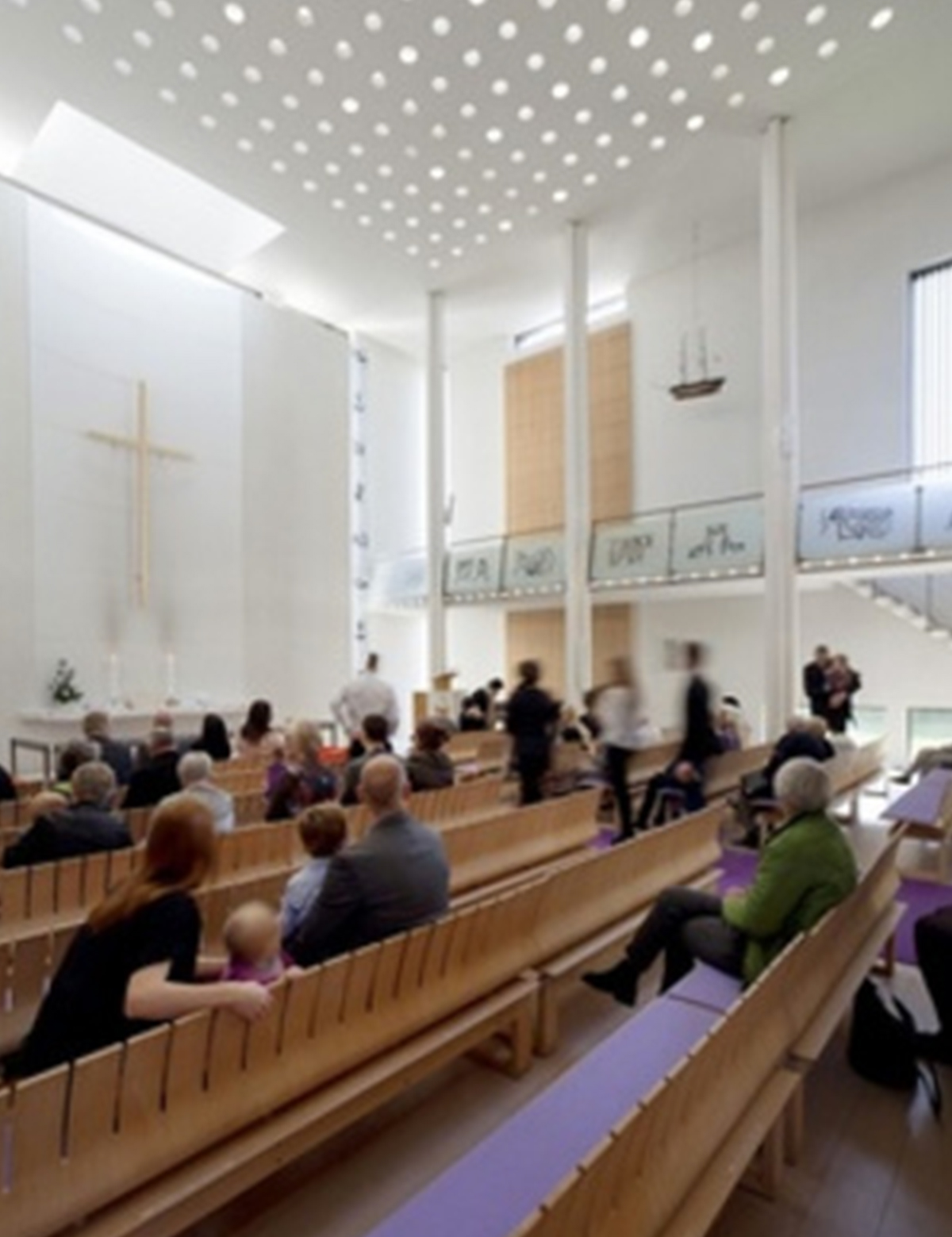 Bredt billede af et moderne kirkeinteriør med højt, hvidt loft med perforeringer, en forsamling siddende i træbænke med lilla hynder og et kors på altervæggen.