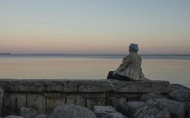 En kvinde i jakke og baret sidder på en lav betonmur ved vandet og kigger ud over havet ved solnedgang eller daggry.