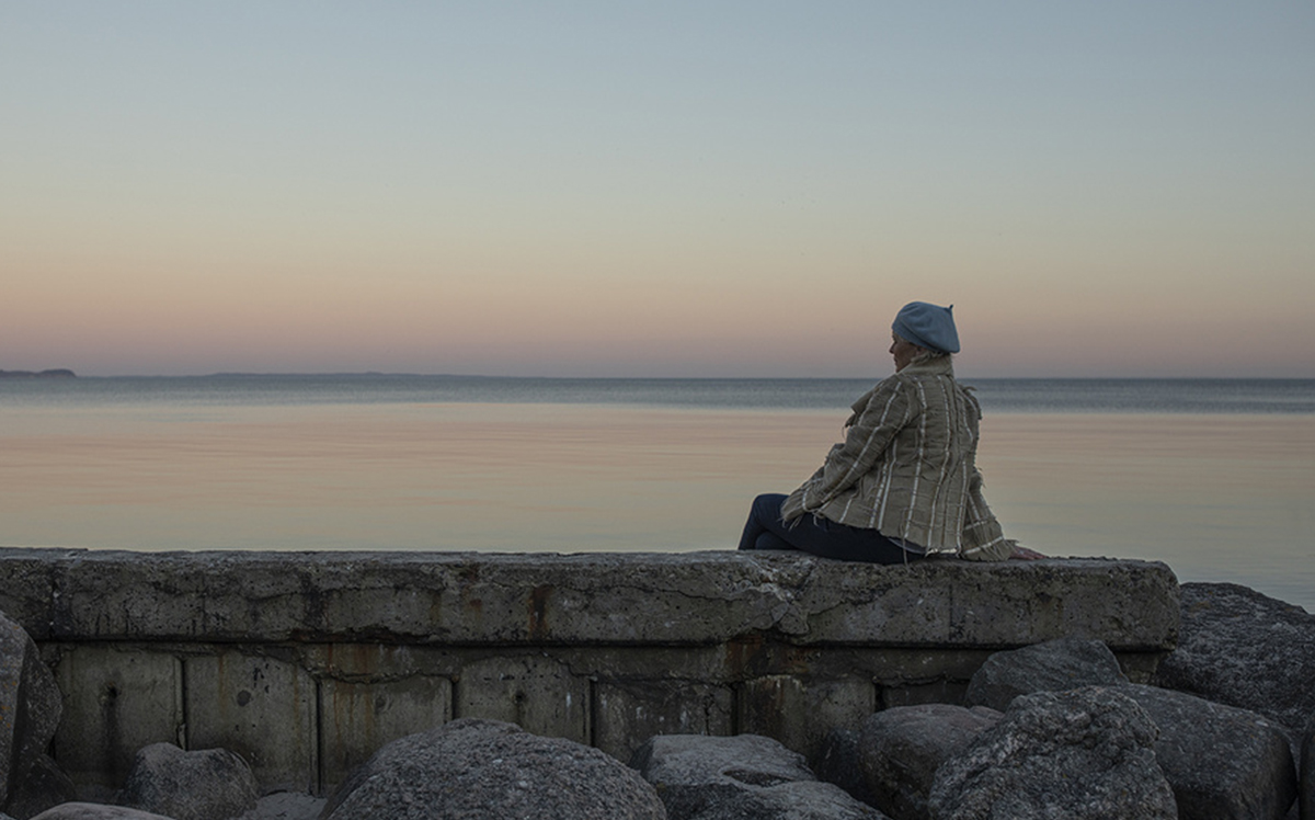 En kvinde i jakke og baret sidder på en lav betonmur ved vandet og kigger ud over havet ved solnedgang eller daggry.