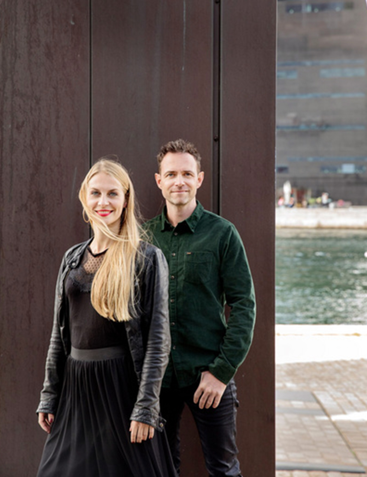 Portræt af en mand og en kvinde, der står side om side foran en mørkebrun væg, med vand og en moderne bygning i baggrunden.