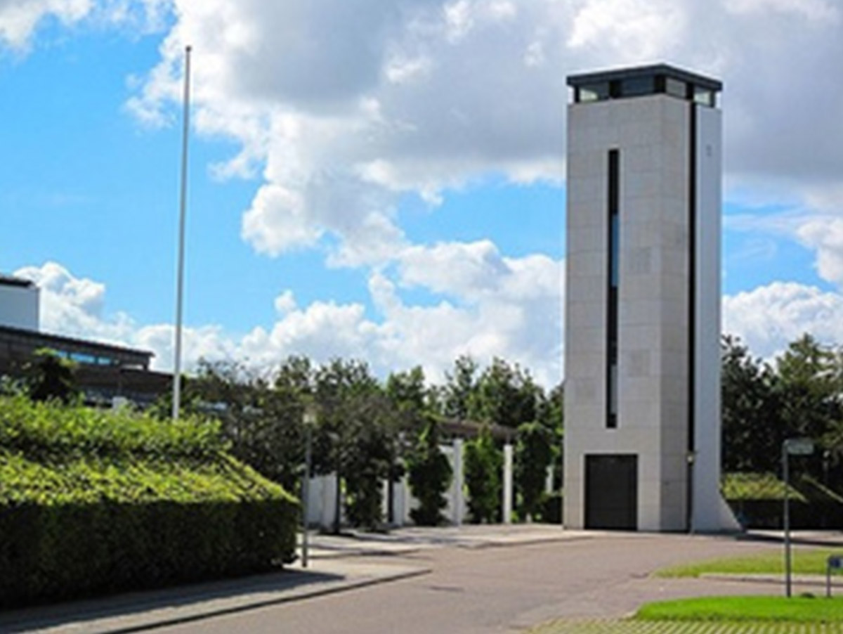 Et moderne fritstående klokketårn i lys sten med et højt, smalt vindue, placeret ved en vej og omgivet af hække og træer under en blå himmel med skyer.