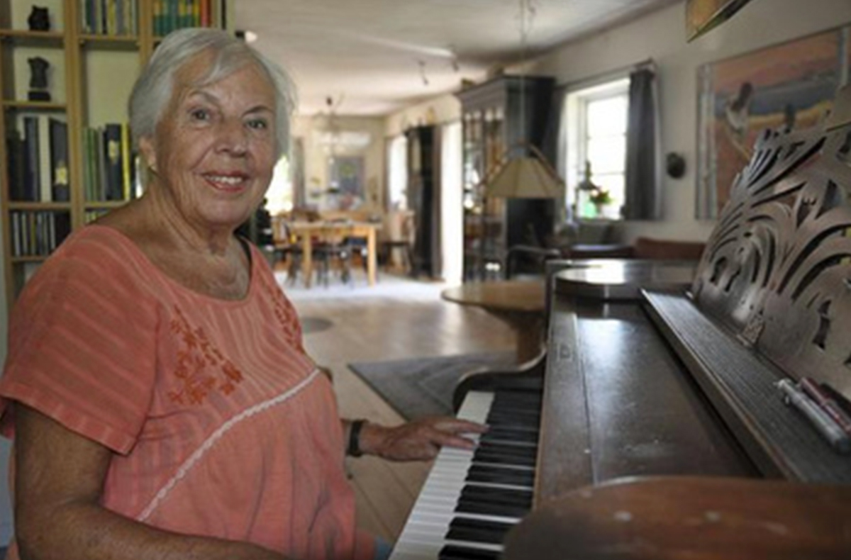 Portræt af en smilende ældre kvinde med gråt hår, der spiller på et træklaver i et hyggeligt, velmøbleret rum med bogreoler og malerier.