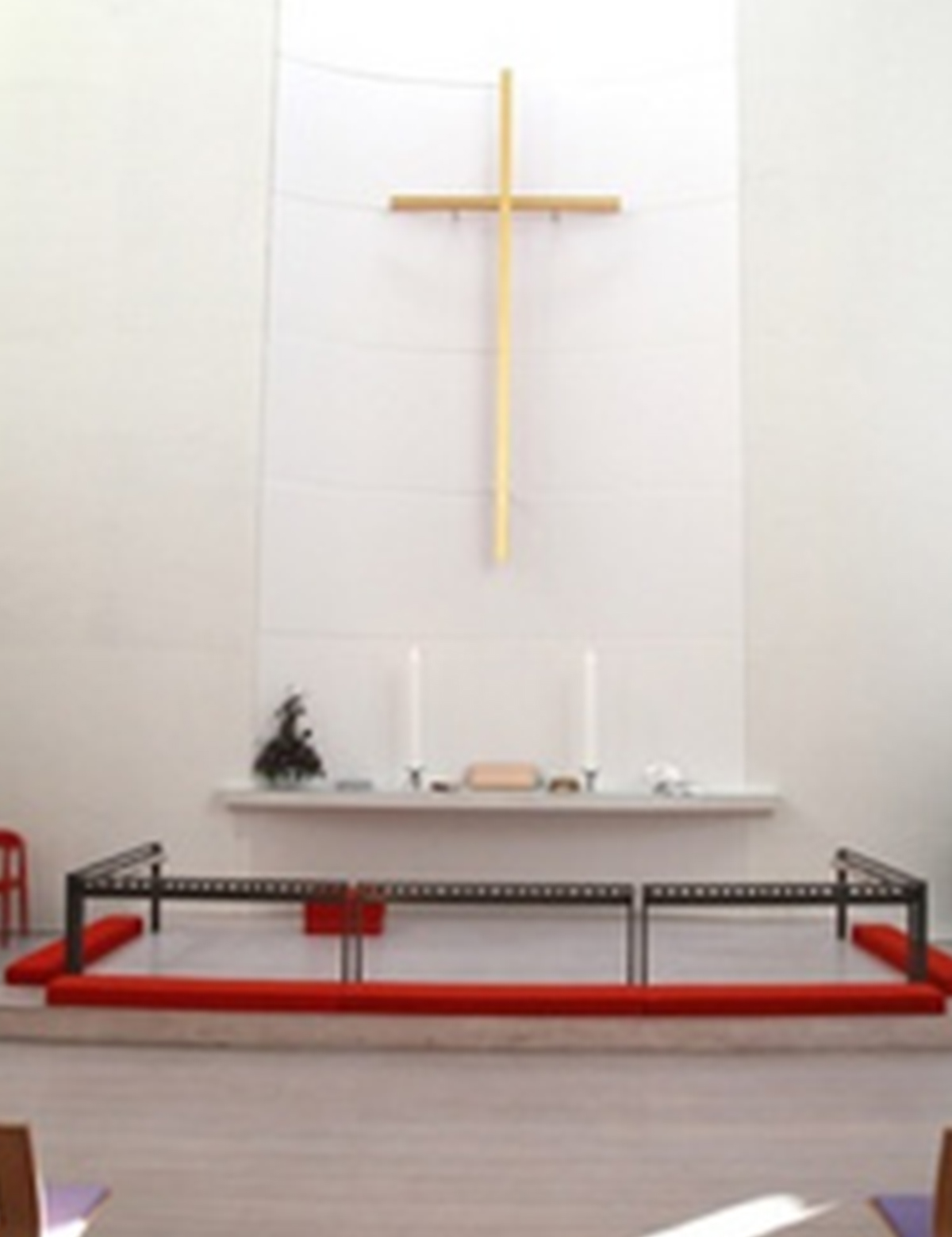 Nærbillede af et moderne alter med et simpelt trækors på en hvid væg, to høje lys, og et rødt tæppeområde med et lavt knæfald.