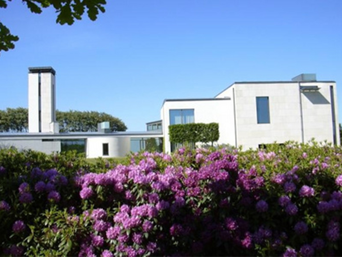 Moderne bygning i lys sten med højt tårn bag en hæk af lilla rododendroner under blå himmel.