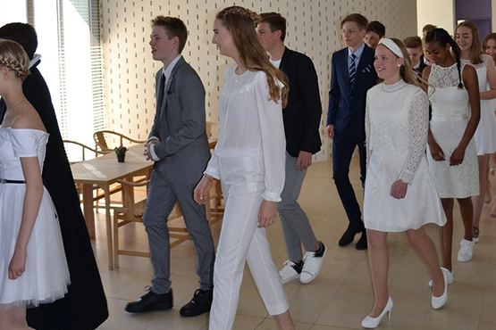 En gruppe teenagere, klædt i festtøj, hovedsageligt hvidt og sort/gråt, går i en linje indendørs, muligvis til en konfirmation eller anden ceremoni.