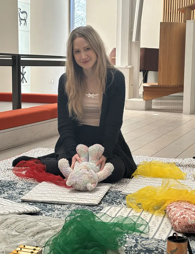 Portræt af en ung kvinde med langt blondt hår, der sidder på et tæppe i en kirke med en bamse og farvede tørklæder.