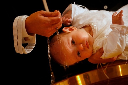 Nærbillede af et spædbarn i hvid dåbskjole, som får hældt vand over panden under en dåbsceremoni.