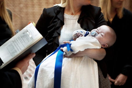 Nærbillede af en baby, der sover i en hvid dåbskjole med et blåt bånd, holdt af en kvinde, mens en person i forgrunden holder en bog.