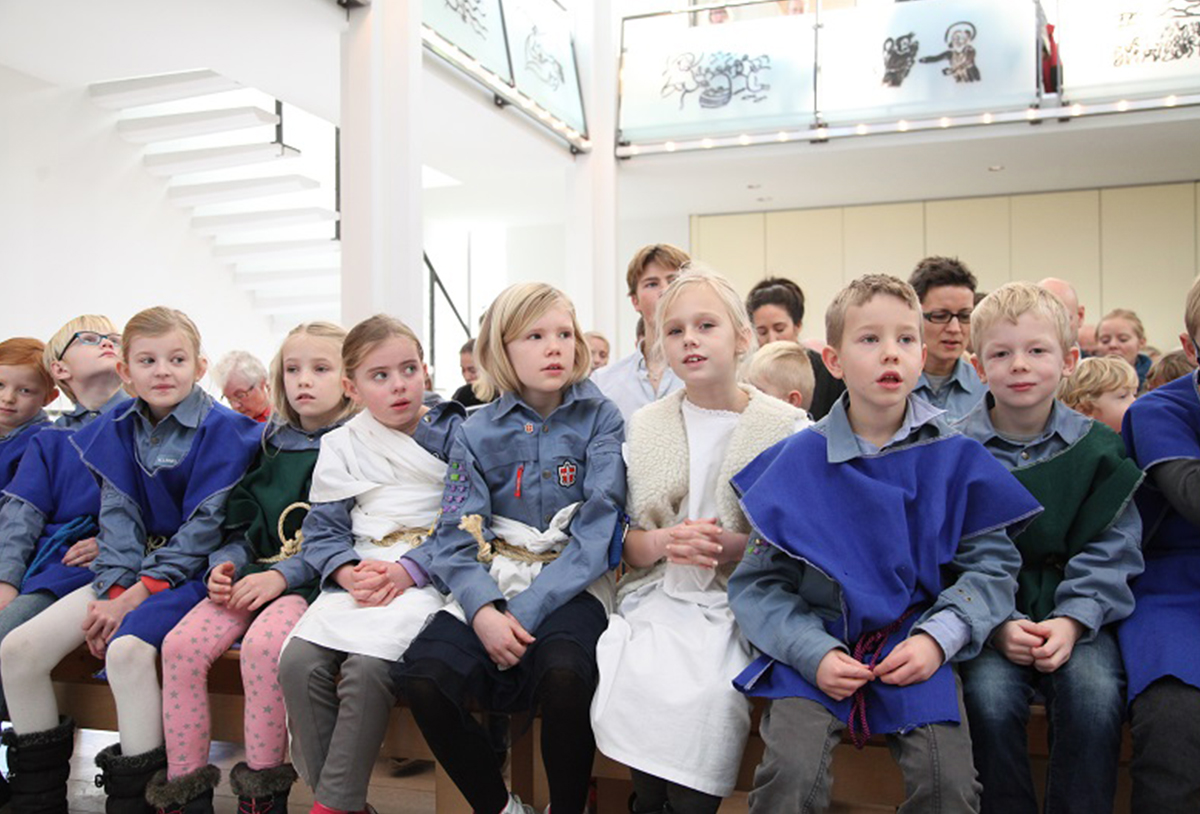 En gruppe børn i forskellige kostumer, primært blå kapper og skjorter, sidder på en bænk i et lyst rum med en hvid trappe i baggrunden.